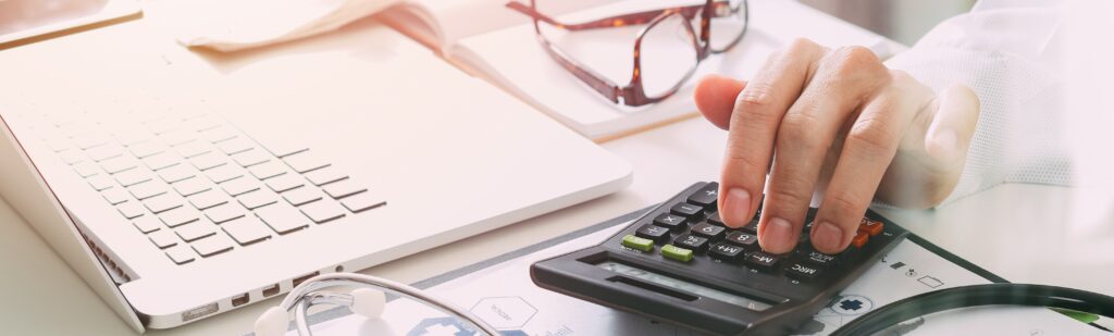 A person uses a calculator on a desk with a laptop, glasses, and a stethoscope nearby, hinting at healthcare finances or medical billing, possibly leveraging APCD data.