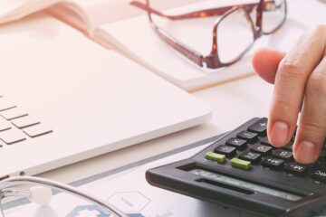 A person uses a calculator on a desk with a laptop, glasses, and a stethoscope nearby, hinting at healthcare finances or medical billing, possibly leveraging APCD data.