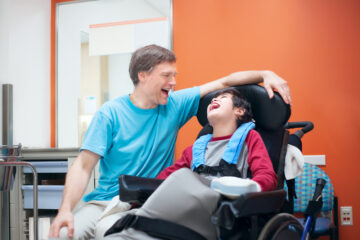 Child in wheelchair laughing with male adult sitting on male child's right side.