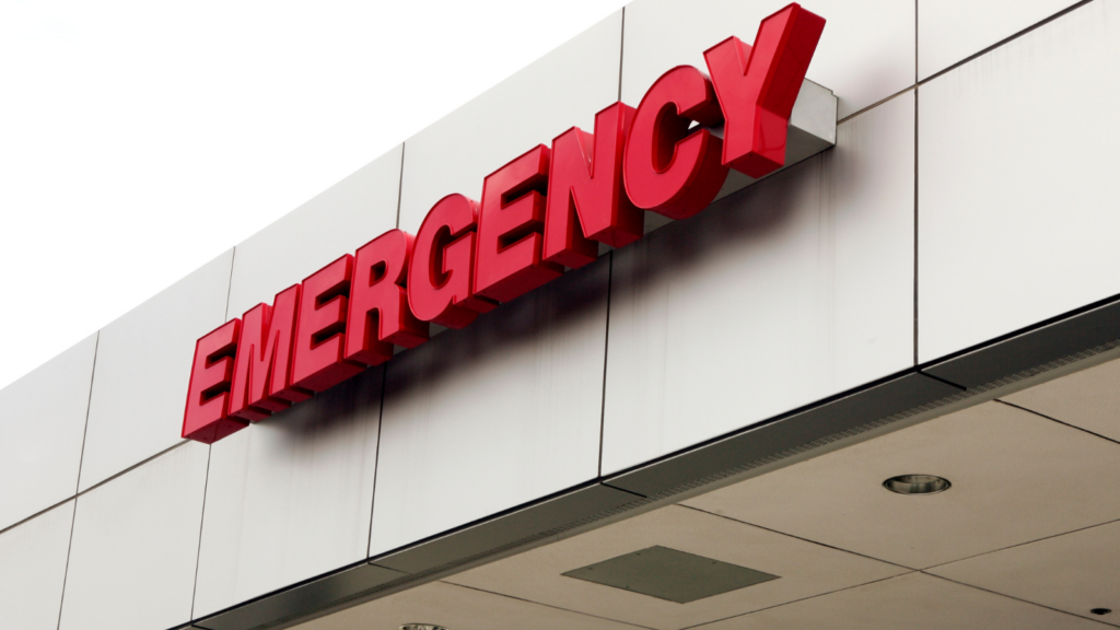 Stock image of a hospital emergency department entrance, with illuminated signage and sliding glass doors.