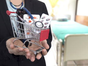 Person in a black clothing, with a hospital bed in the background is wearing a stethoscope and holding with both hands a miniature shopping cart with a variety of vaccines, medicines placed inside the cart.